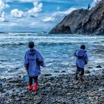 Photo of two children wearing rain jackets, boots on rocky beach by water